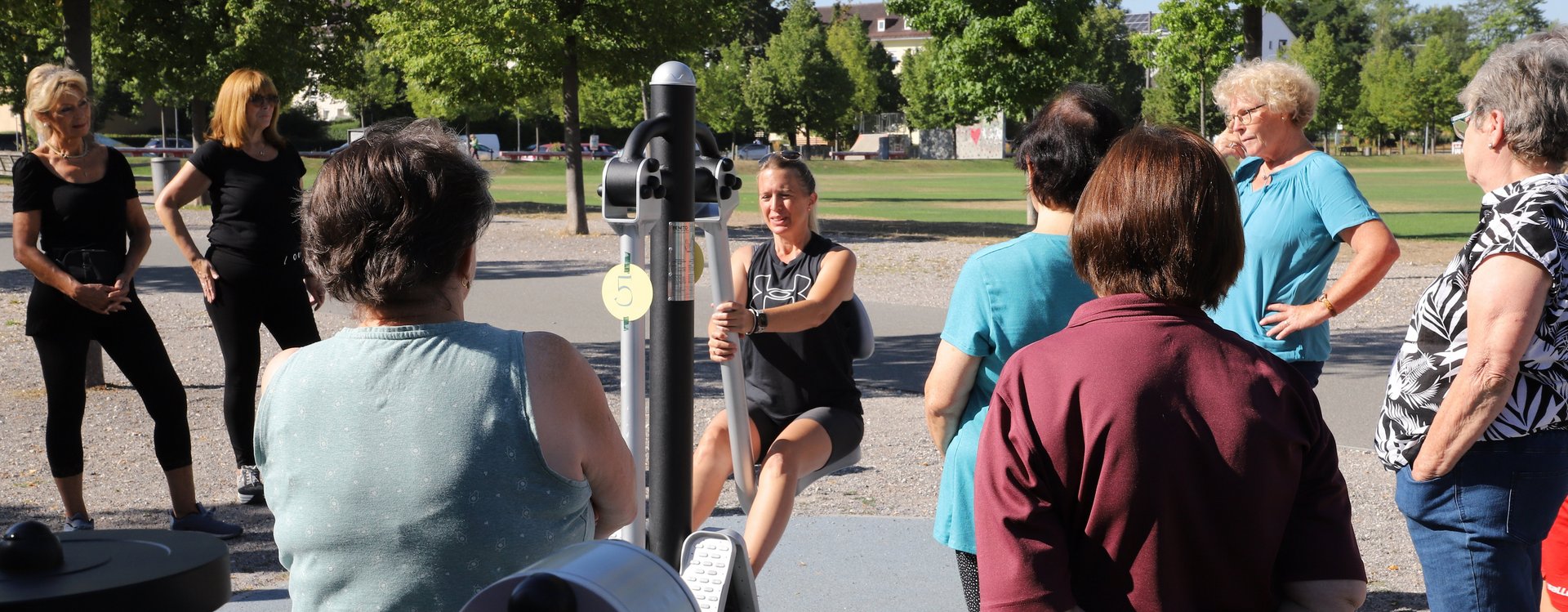 Eine Trainerin demonstriert einer Gruppe von Seniorinnen die korrekte Ausführung eines Fitnessgeräts.