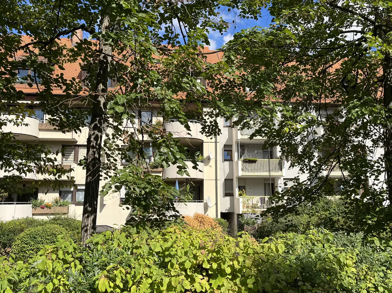 Blick auf die Fassade mit Balkonen von mehrstöckigen Mietshäusern. Im Vordergrund stehen Hecken und Bäume.