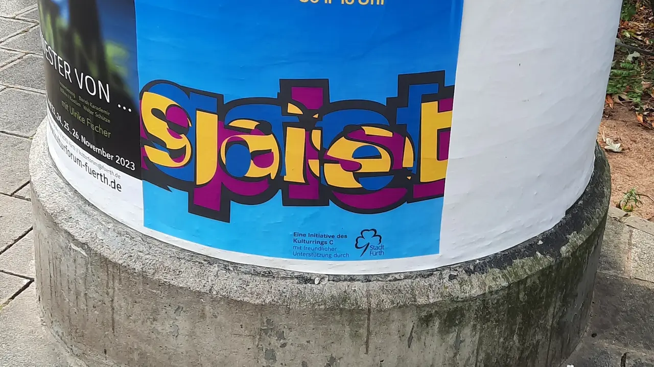 An einer Litfassäule hängt das Plakat mit dem Gastspiellogo: Das Wort "Gastspiel" mit eineinander verschränkten Buchstaben.