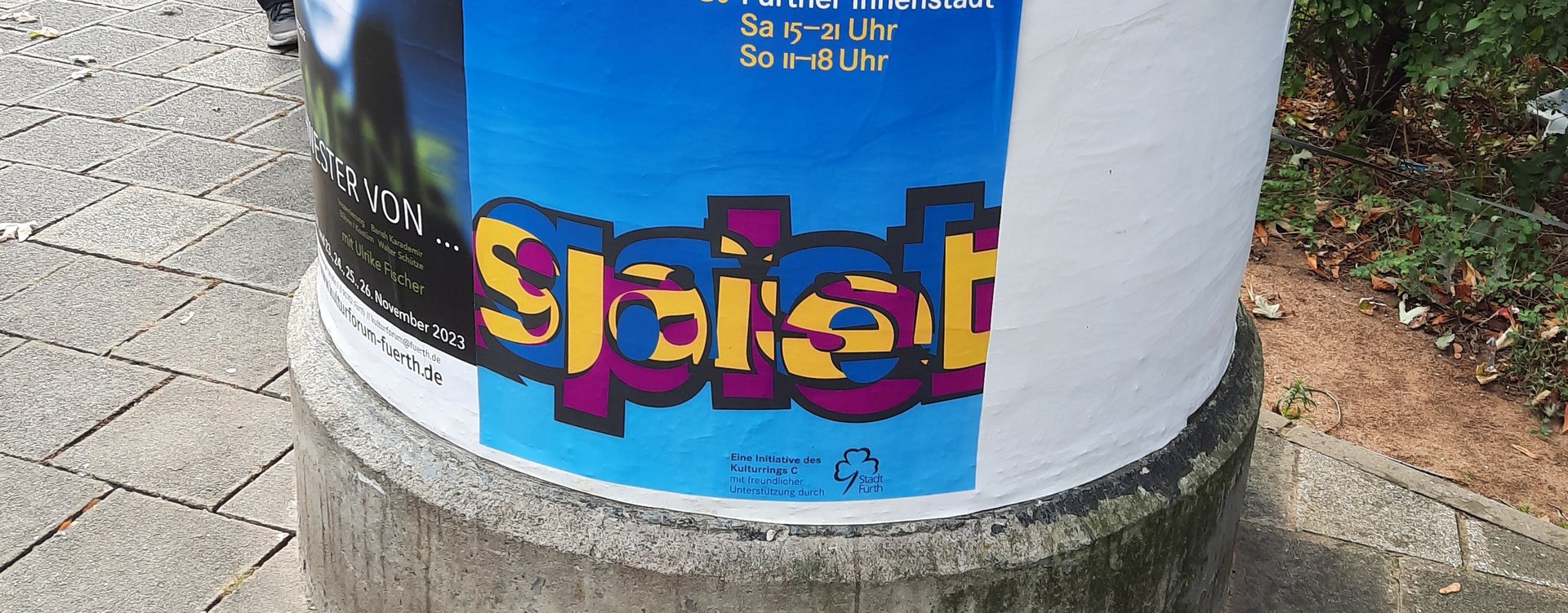 An einer Litfassäule hängt das Plakat mit dem Gastspiellogo: Das Wort "Gastspiel" mit eineinander verschränkten Buchstaben.