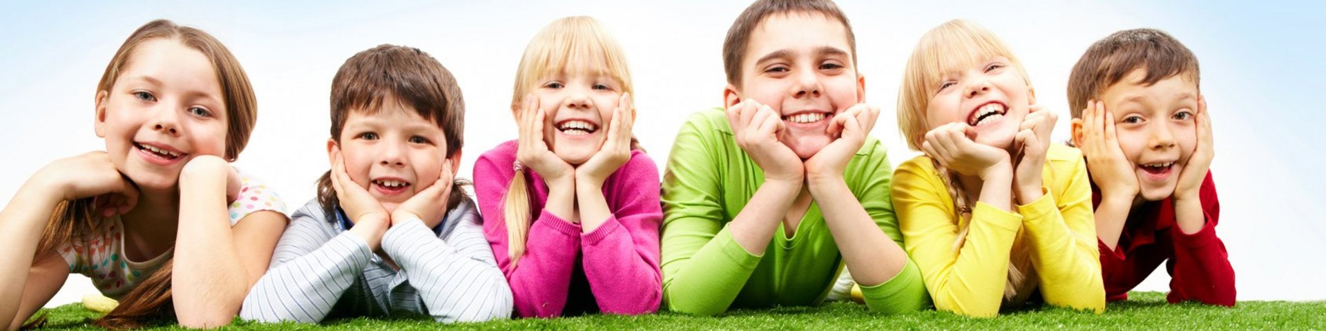 Sechs Kinder liegen bäuchlings auf einer Wiese, das Gesicht auf die Hände gestützt, lachend.