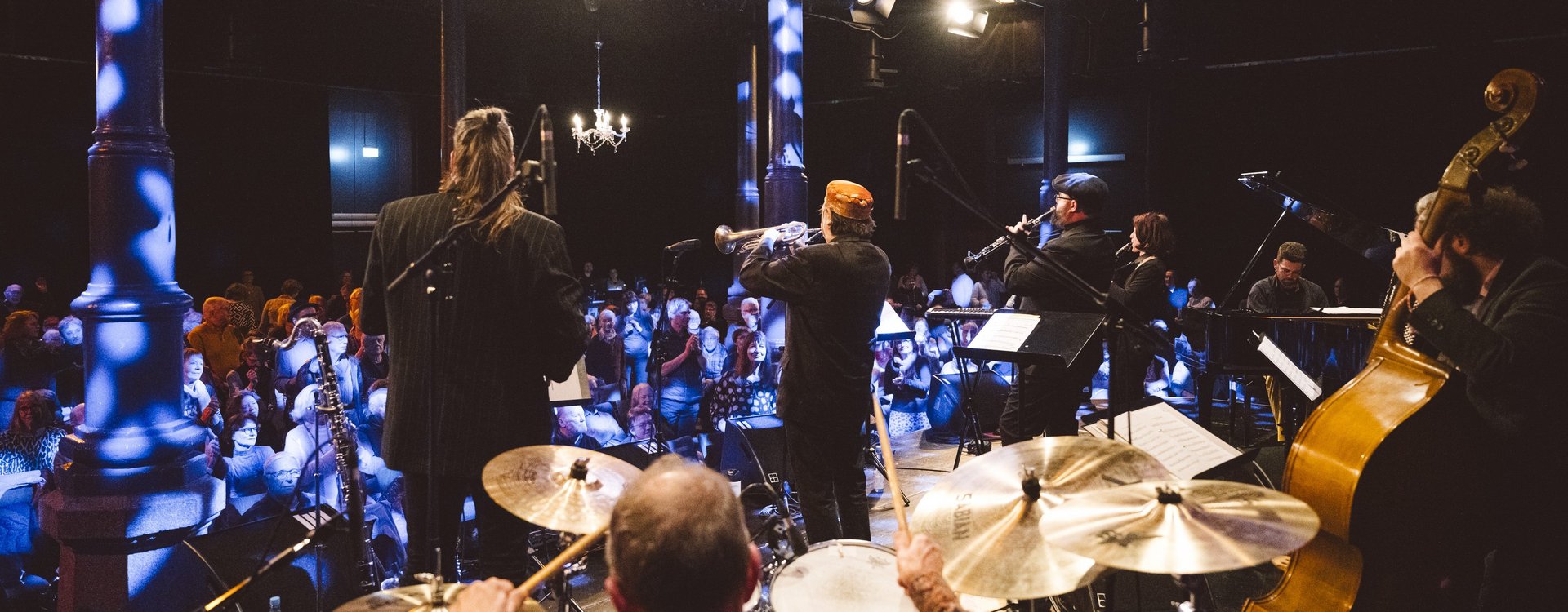 Konzert im Kulturforum aus der Bühnenperspektive fotografiert. Im Vordergrund Schlagzeug, Kontrabass und Bläser, dahinter das Publikum.