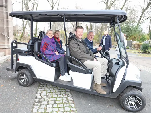 Der Oberbürgermeister sitzt am Steuer des weißen Friedhofmobils, neben ihm ein Mann, auf den hinteren beiden Plätzen sitzen zwei Frauen. Klick öffnet Bild in größerer Ansicht.