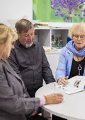 Eine Frau in einem fliederfarbenem Jacket und ein älteres Paar sitzen an einem Schreibtisch im Büro, die Frau zeigt ihnen eine Broschüre