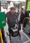 Eine ältere Dame steigt mit ihrem Rollator in einen Bus ein, ein Mann gibt ihr dabei Tipps