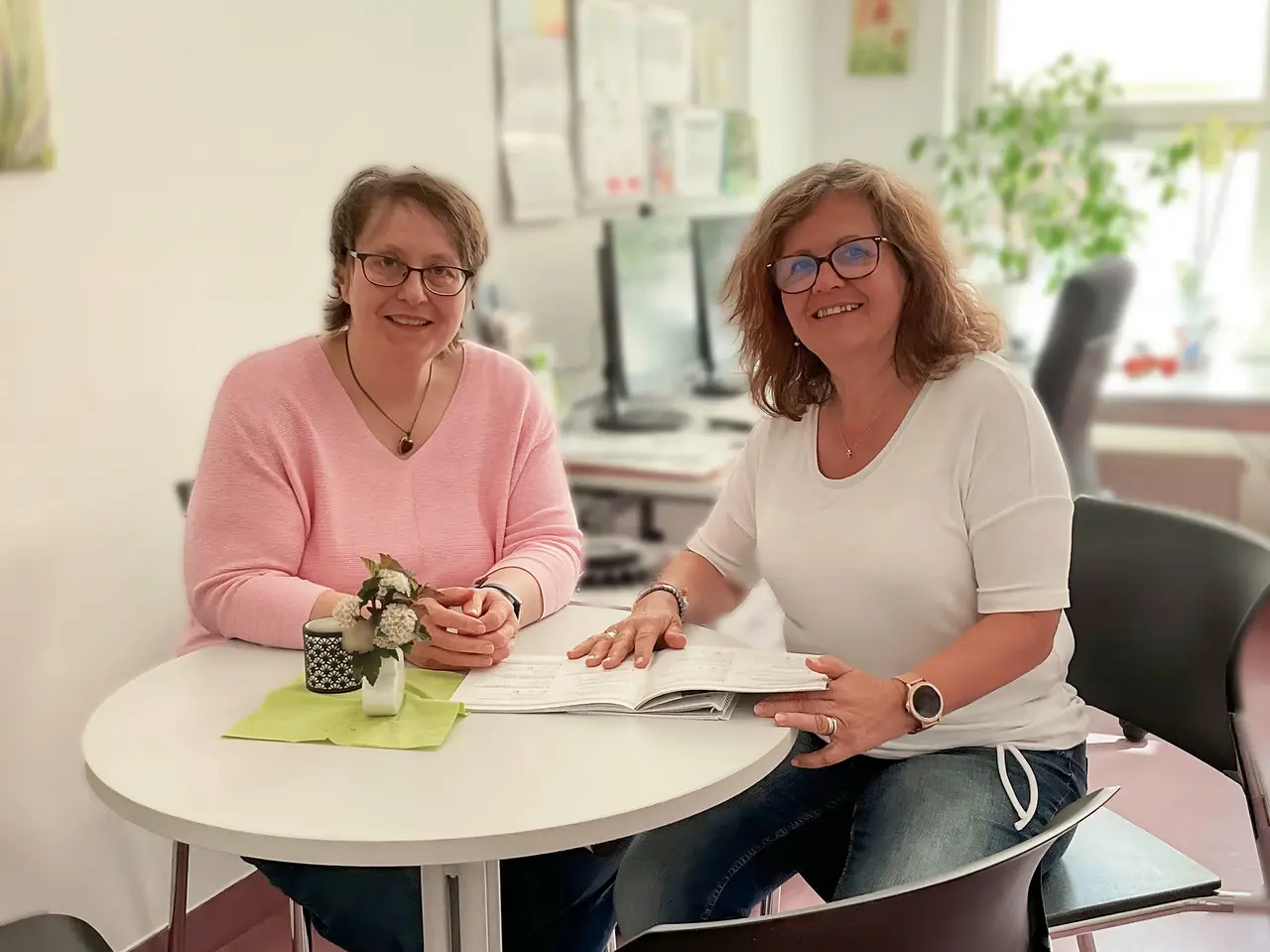 Zwei Frauen sitzen an einem runden Tisch in einem Büro, eine Broschüre liegt aufgeschlagen in der Mitte.