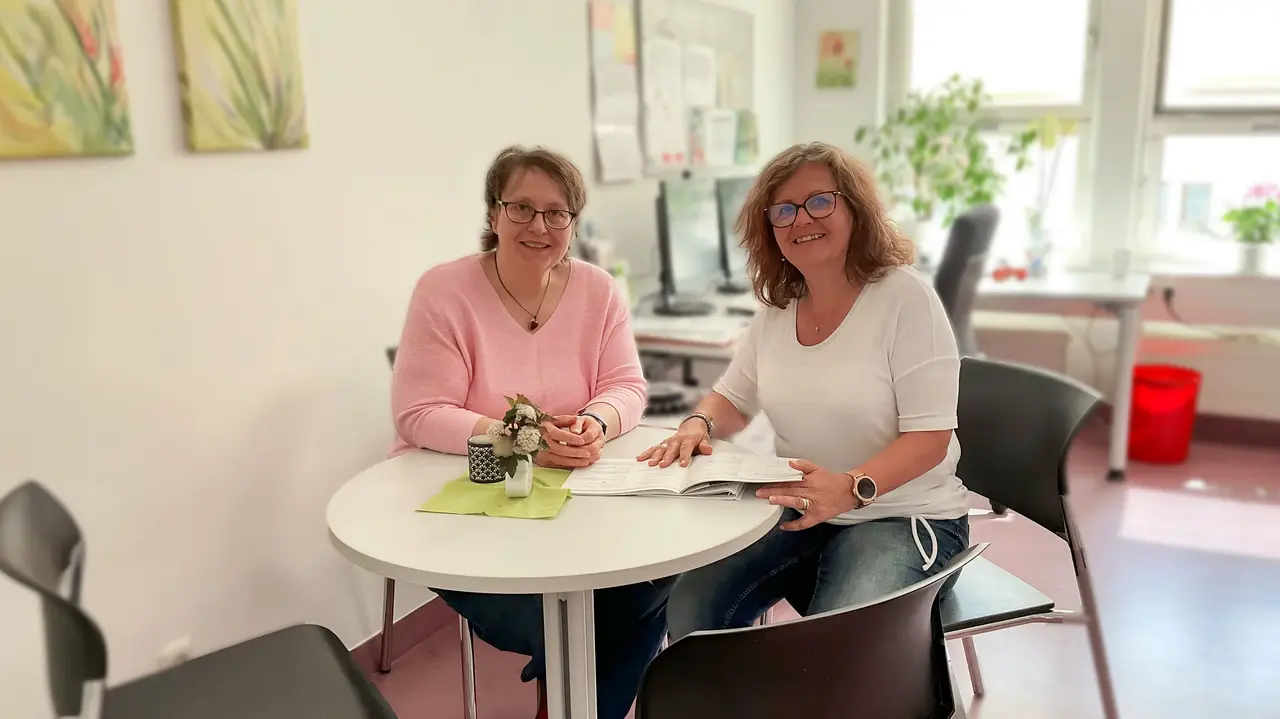 Zwei Frauen sitzen an einem runden Tisch in einem Büro, eine Broschüre liegt aufgeschlagen in der Mitte.