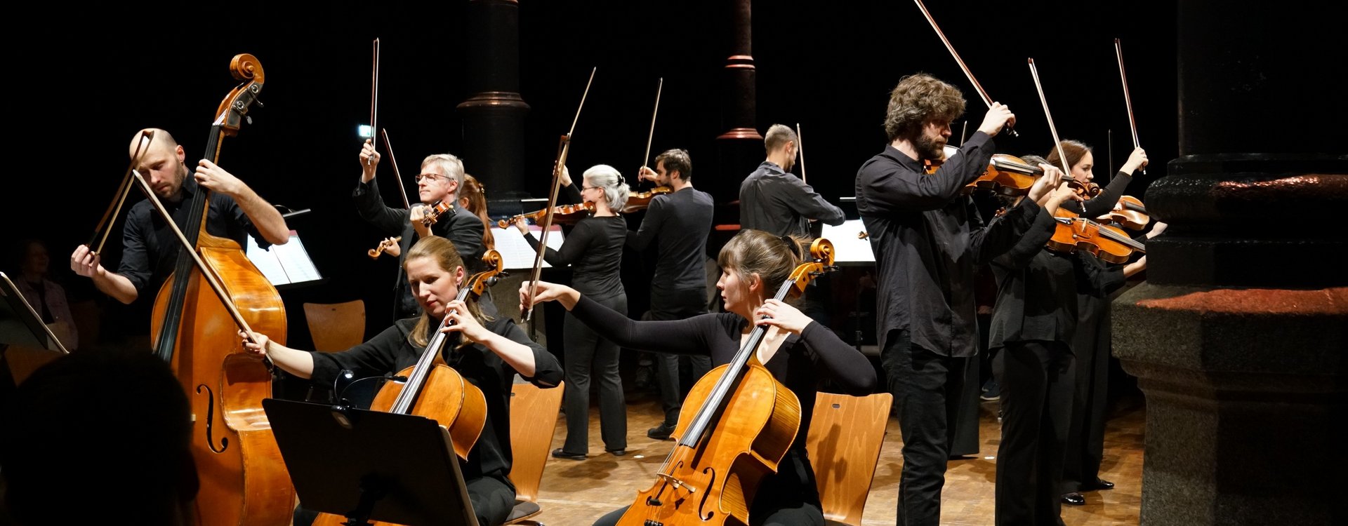 Das Orchester Venturo steht im Kreis im großen Saal des Kulurforums mit den markanten Säulen und musiziert.