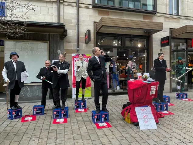 Männer und Frauen in Anzügen stehen auf Getränkekästen in der Fußgängerzone, um sie herum verschiedene Poster zum Equal Pay Day. Klick öffnet Bild in größerer Ansicht.