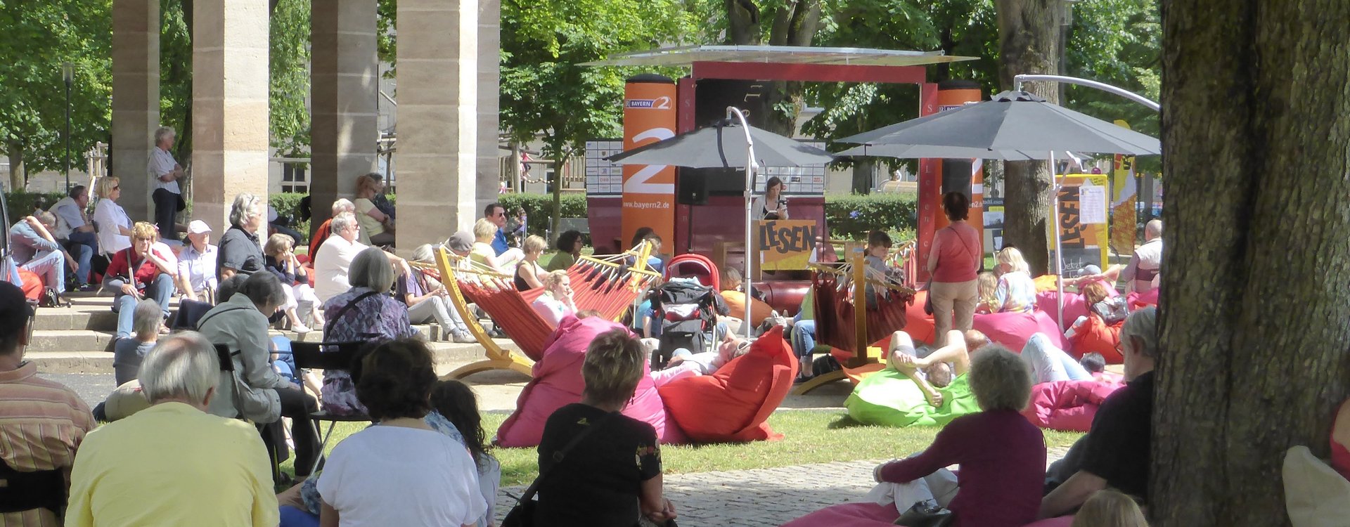 Auf der Adenaueranlage sitzt Publikum bei strahlendem Sonnenschein auf mitgebrachten Sitzgelegenheiten, um einer Lesung auf der Bühne beizuwohnen.