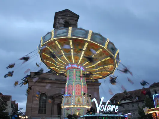 Fahrgeschäft "Volare" beleuchtet vor Kirche und dämmerndem Himmel; Klick öffnet Bild in größerer Ansicht.