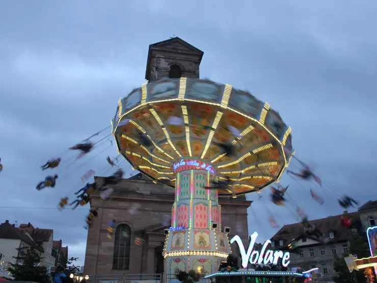 Fahrgeschäft "Volare" beleuchtet vor Kirche und dämmerndem Himmel; Klick öffnet Bild in größerer Ansicht.