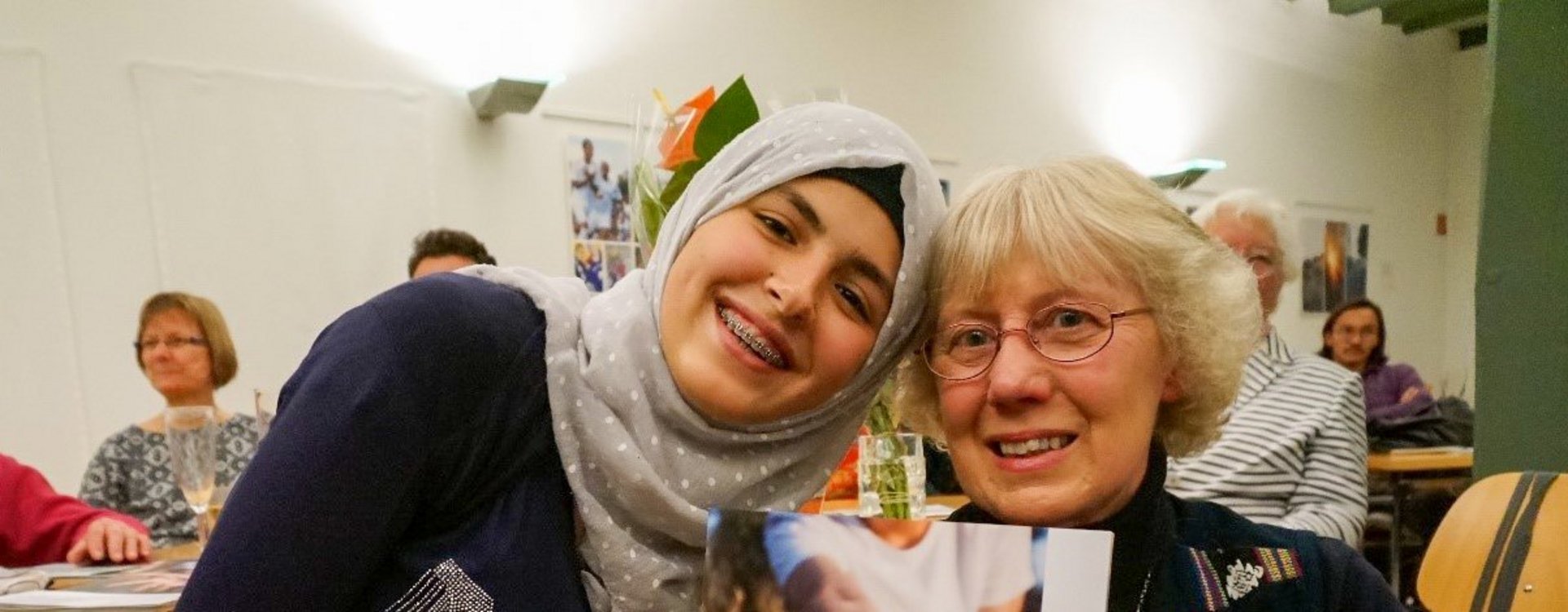 Eine junge Frau mit Kopftuch und eine ältere Frau halten gemeinsam ein Foto mit einer offenen Hand, auf der "Wir helfen" steht, in die Kamera.