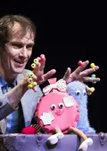 Ein Schauspieler spielt mit bunten Finger- und Handpuppen, die auf einem alten Koffer sitzen.