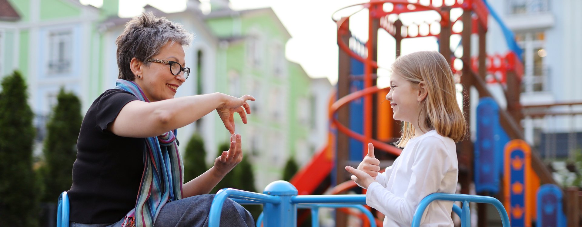 Eine Frau kommuniziert in Gebärdensprache mit einem Mädchen während sie auf einem Spielplatzkarussell sitzen.