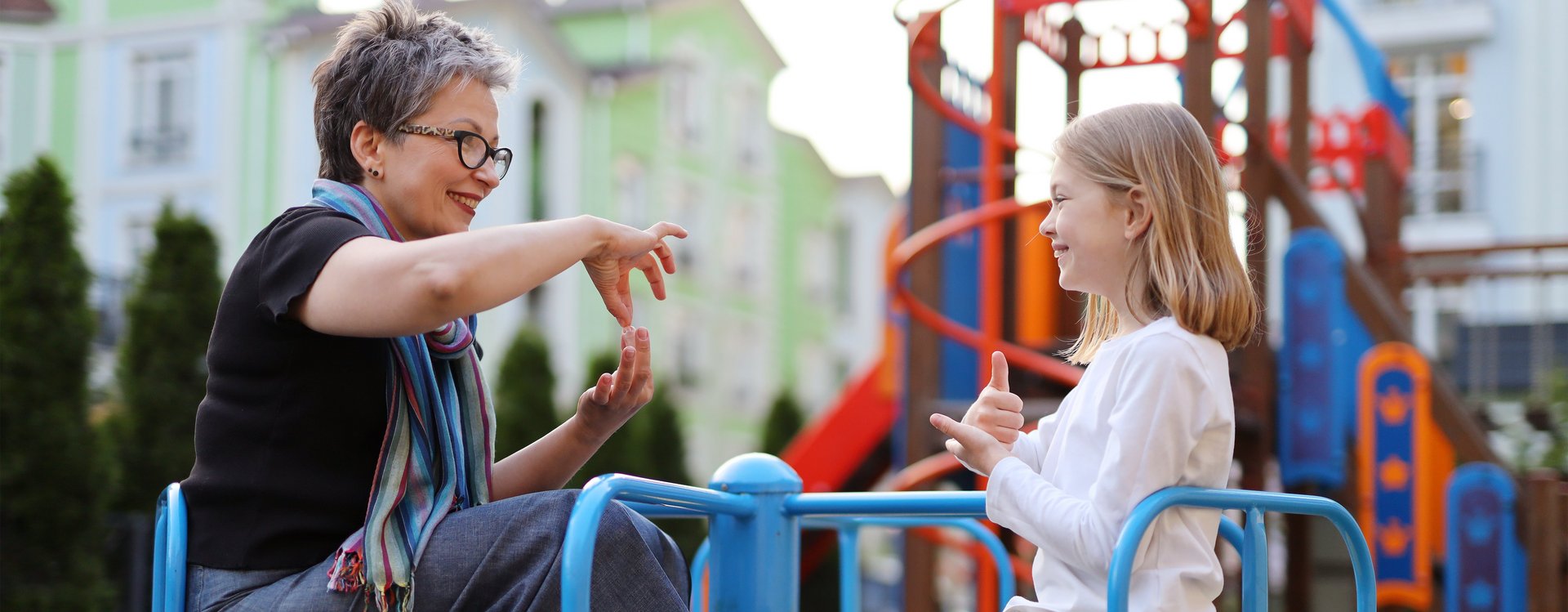 Eine Frau kommuniziert in Gebärdensprache mit einem Mädchen während sie auf einem Spielplatzkarussell sitzen.