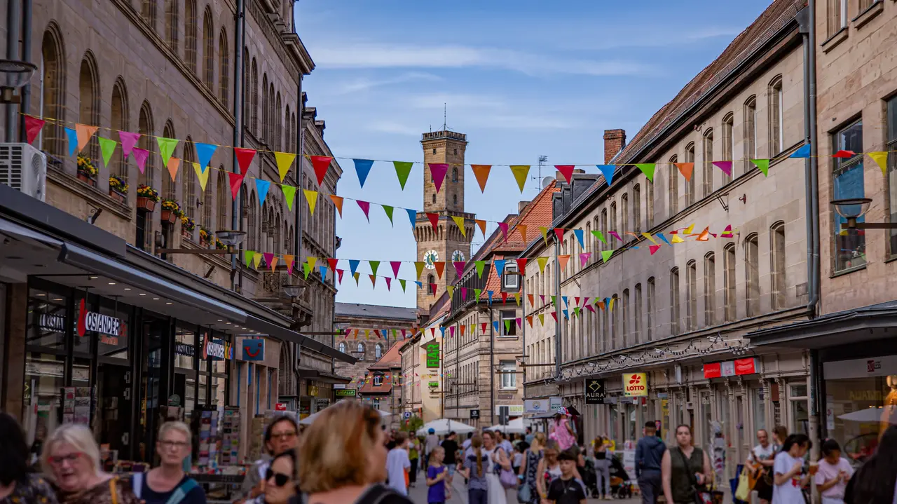 Die belebte Fußgängerzone mit dem Rathausturm im Hintergrund. Bunte Wimpel sind über die Straße gepannt.