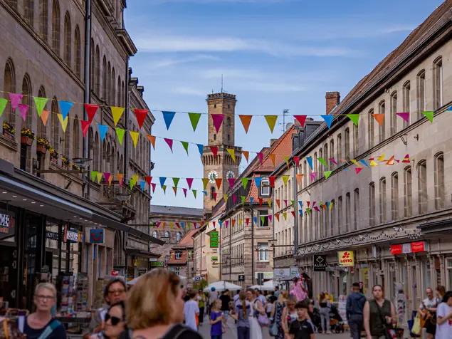 Die belebte Fußgängerzone mit dem Rathausturm im Hintergrund. Bunte Wimpel sind über die Straße gepannt.