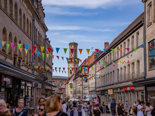 Die belebte Fußgängerzone mit dem Rathausturm im Hintergrund. Bunte Wimpel sind über die Straße gepannt.