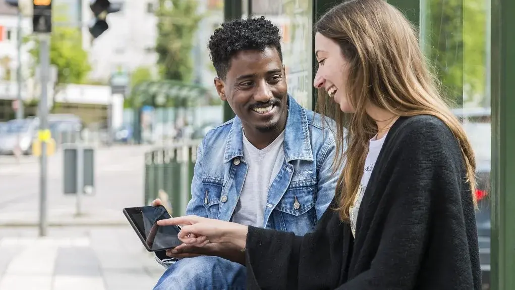 Ein Mann und eine Frau sitzen an einer Haltestelle, sie zeigt auf sein Tablet